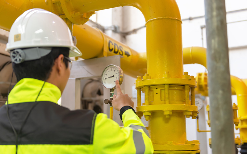 Technician in hardhat and yellow jacket points to a gauge on yellow industrial pipes.