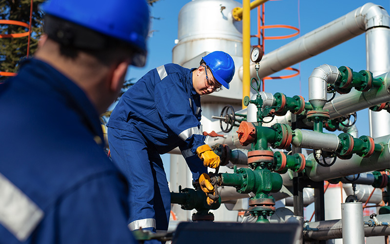Two workers in blue protective gear adjusting valves on industrial piping at a large processing facility.