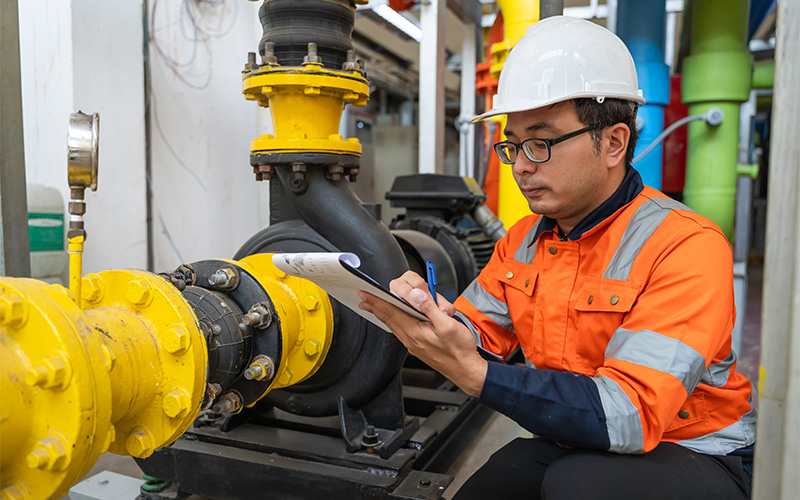 Worker in safety gear inspects machinery, writing notes on clipboard.